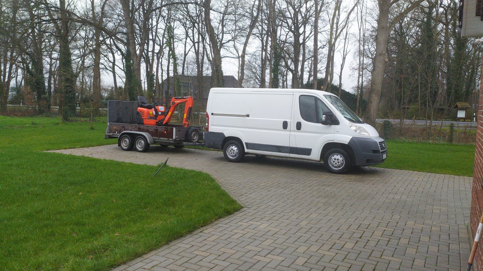 Biały samochód dostawczy z przyczepą, na której przewożona jest mała pomarańczowa koparka, zaparkowany na podjeździe z kostki brukowej przed domem w pochmurny dzień.