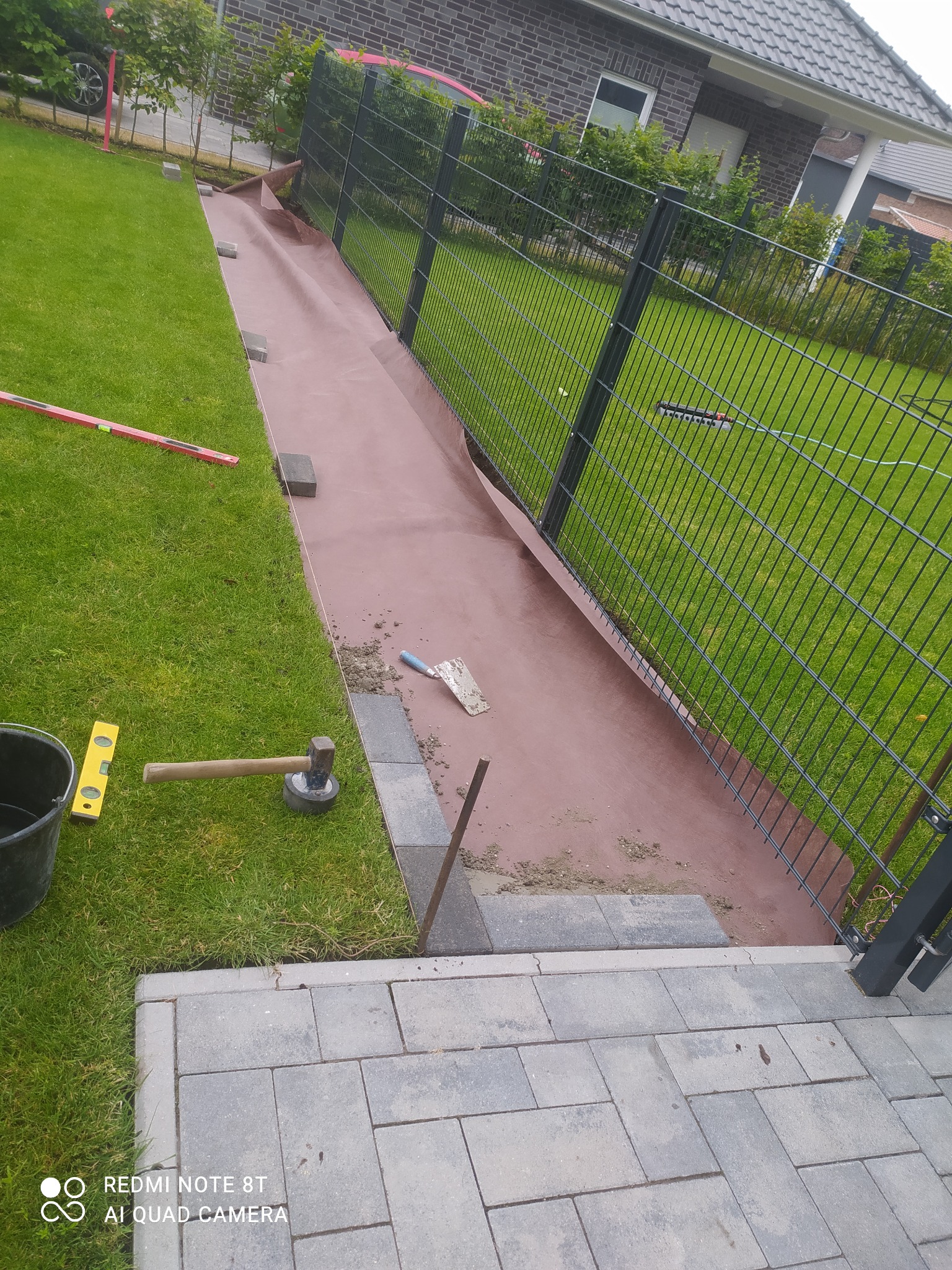 Przygotowanie podłoża pod ścieżkę z kostki brukowej wzdłuż ogrodzenia: rozłożona geowłóknina, obrzeża z kostki, narzędzia pracy (poziomica, młotek, kielnia).