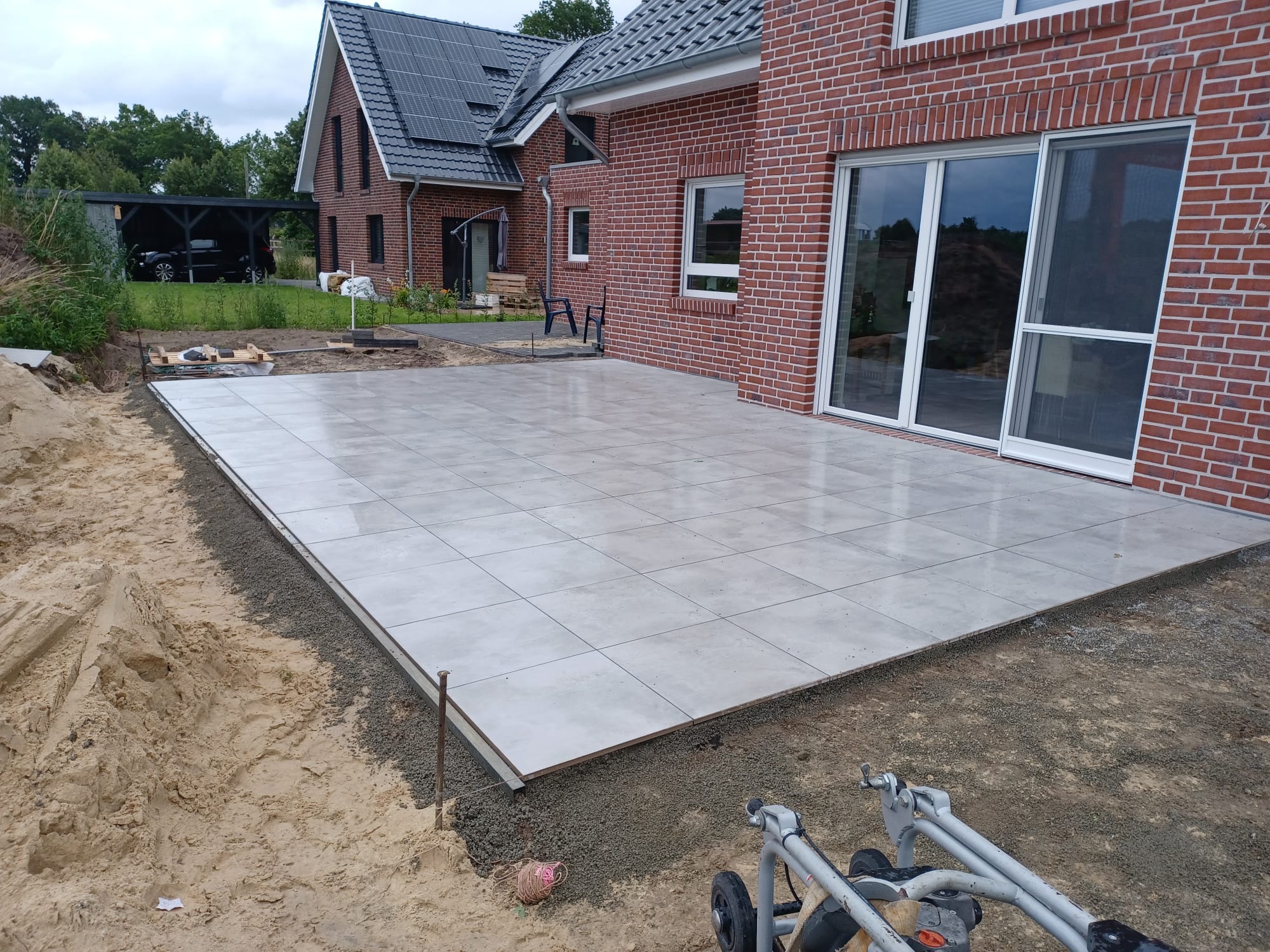Jasnoszara, kwadratowa nawierzchnia z płytek ułożona na zewnątrz, obok ceglanego domu z przesuwnymi drzwiami, widoczny ubijak w tle, etap prac wykończeniowych.