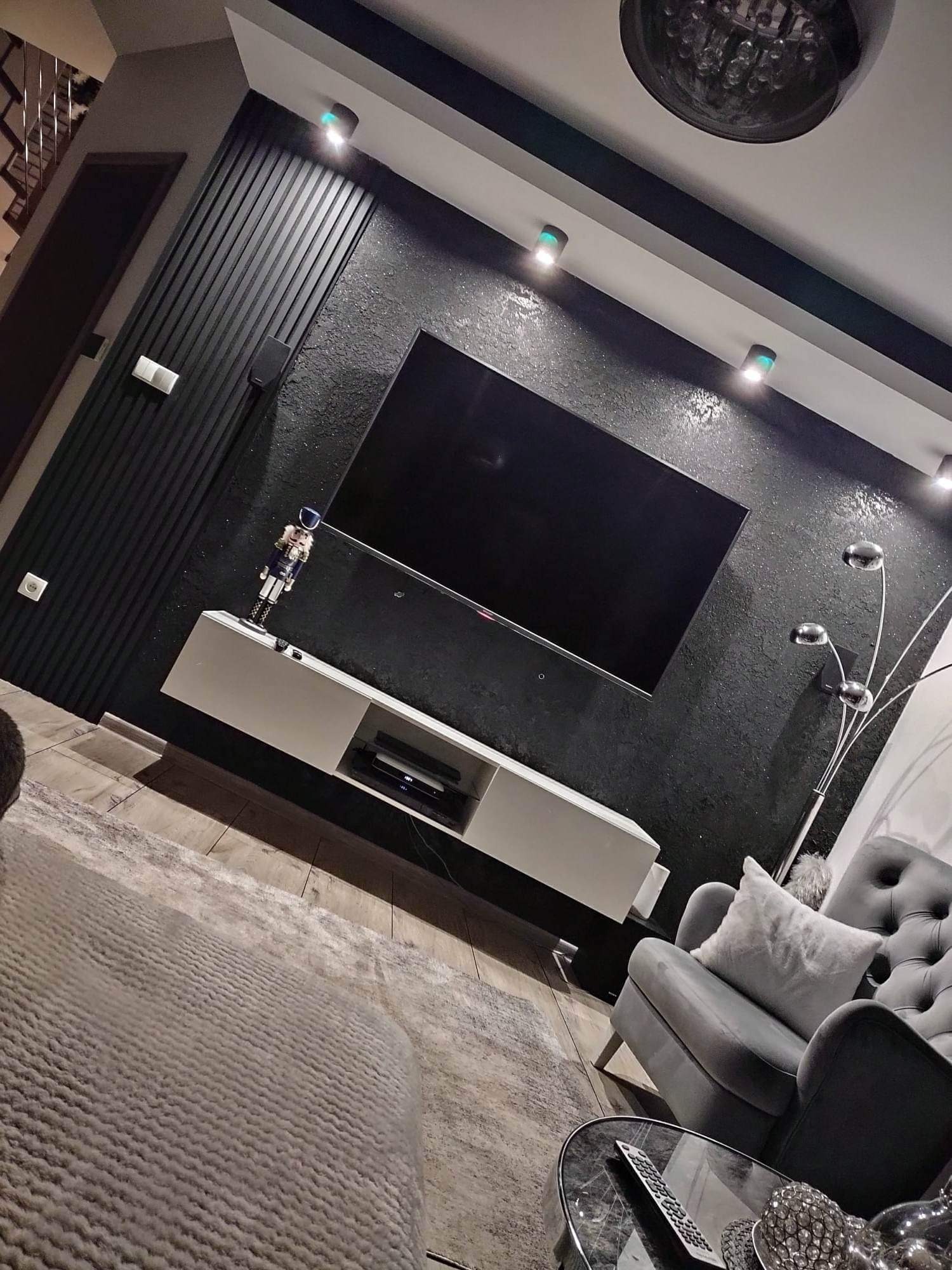 Eleganckie wnętrze salonu z czarną, teksturowaną ścianą, białym, podwieszanym regałem RTV, dużym telewizorem, szarym fotelem typu chesterfield i nowoczesną lampą podłogową. Na regale stoi...