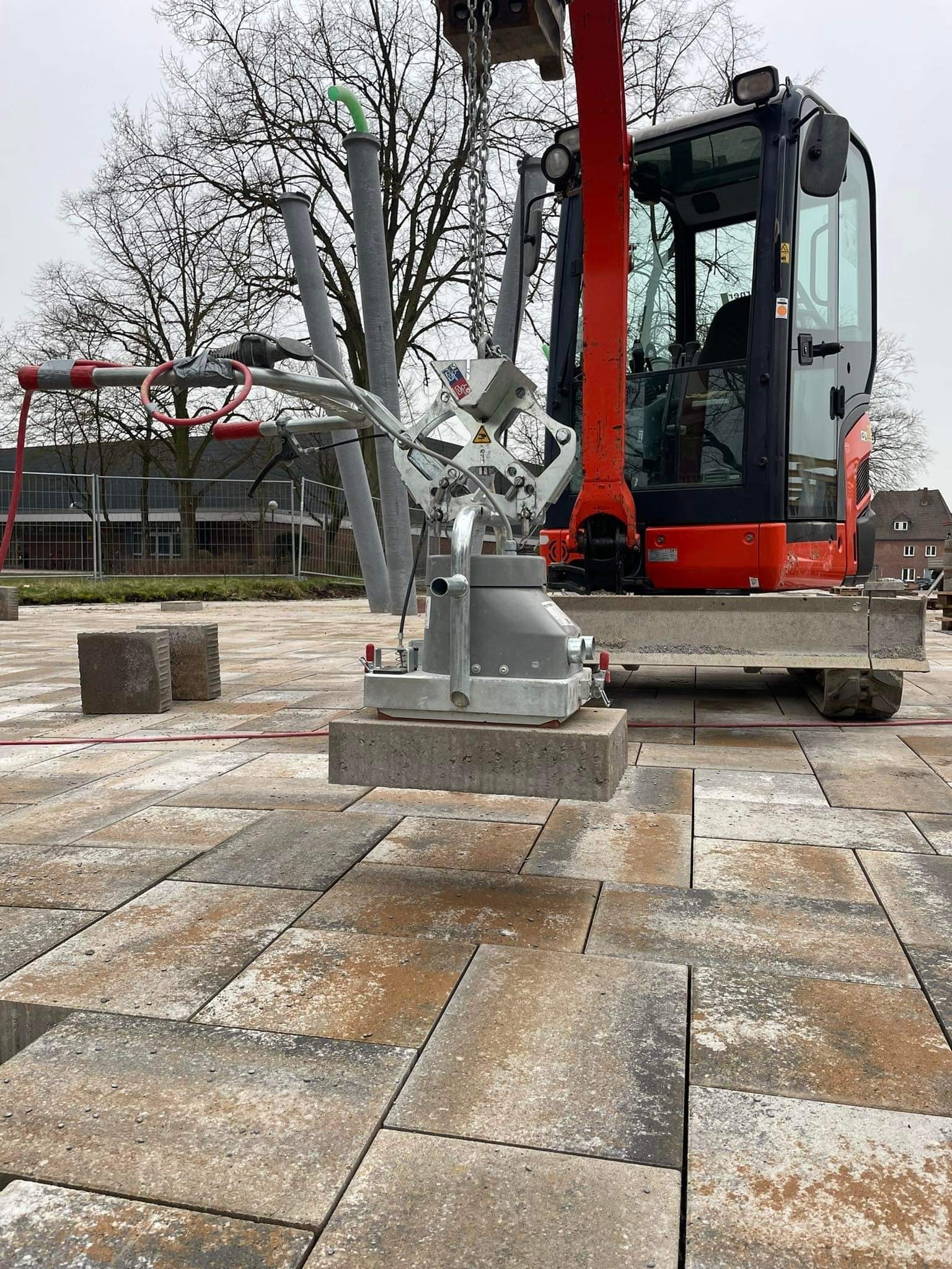 Mini koparka z przyssawką podnosi betonową kostkę brukową na placu budowy, widoczne ułożone już elementy i zapas kostki.