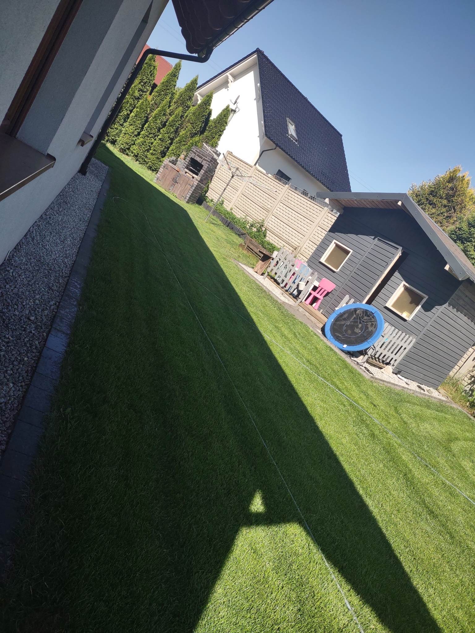 Długi, zielony trawnik z kostką granitową wzdłuż budynku, w tle domek narzędziowy z trampoliną i huśtawką dla dzieci, grill z cegły i wysokie tuje.