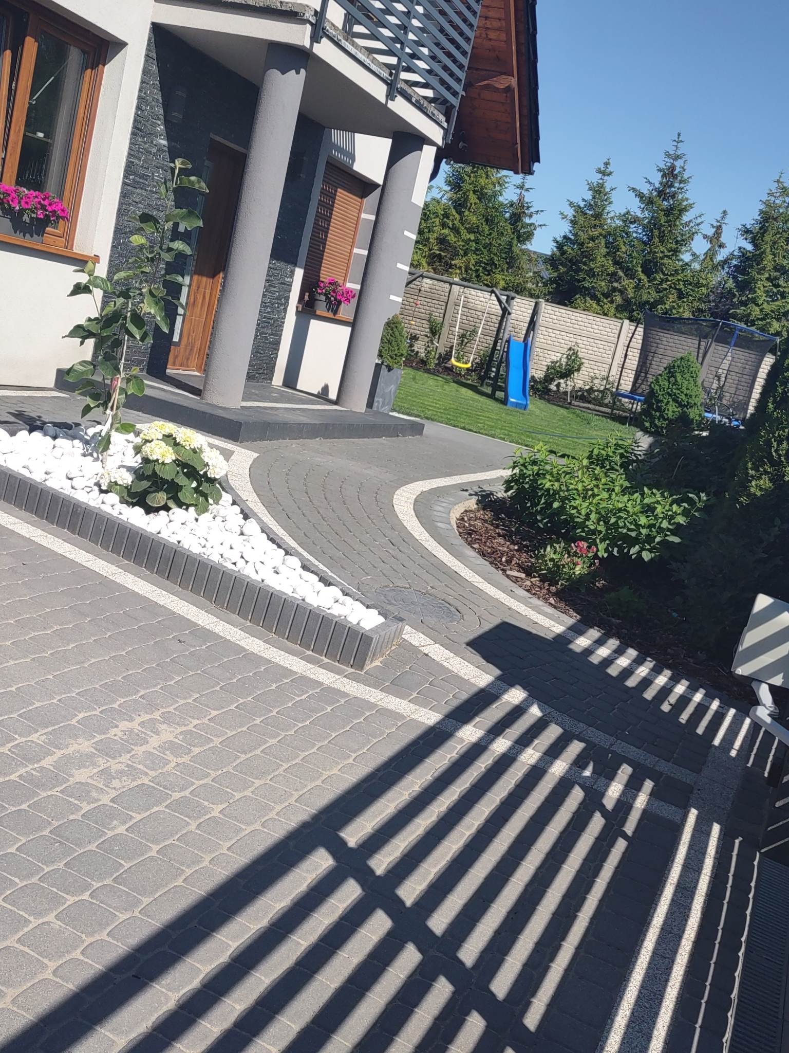 Ułożona kostka brukowa w odcieniach szarości tworząca ścieżkę wokół domu, z białymi kamieniami dekoracyjnymi i zielenią, plac zabaw z huśtawką i zjeżdżalnią w tle.