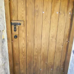 Drewniane drzwi z metalowym zamkiem, widoczne ślady bejcy i faktura drewna, osadzone w kamiennej futrynie.