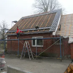 Częściowo pokryty dach z czarnej dachówki, widoczne drewniane łaty, dekarz w czerwonej kurtce na rusztowaniu, ceglany budynek z oknem w tle, pochmurne niebo.