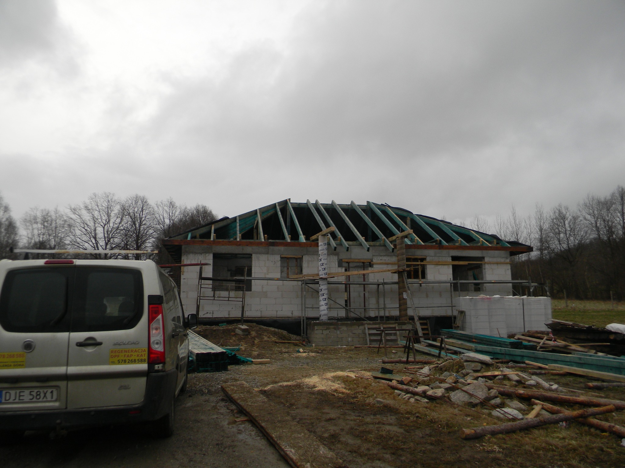 Budowa domu jednorodzinnego w trakcie realizacji, widoczne ściany z bloczków, konstrukcja drewniana dachu przed pokryciem, oraz samochód dostawczy na pierwszym planie.