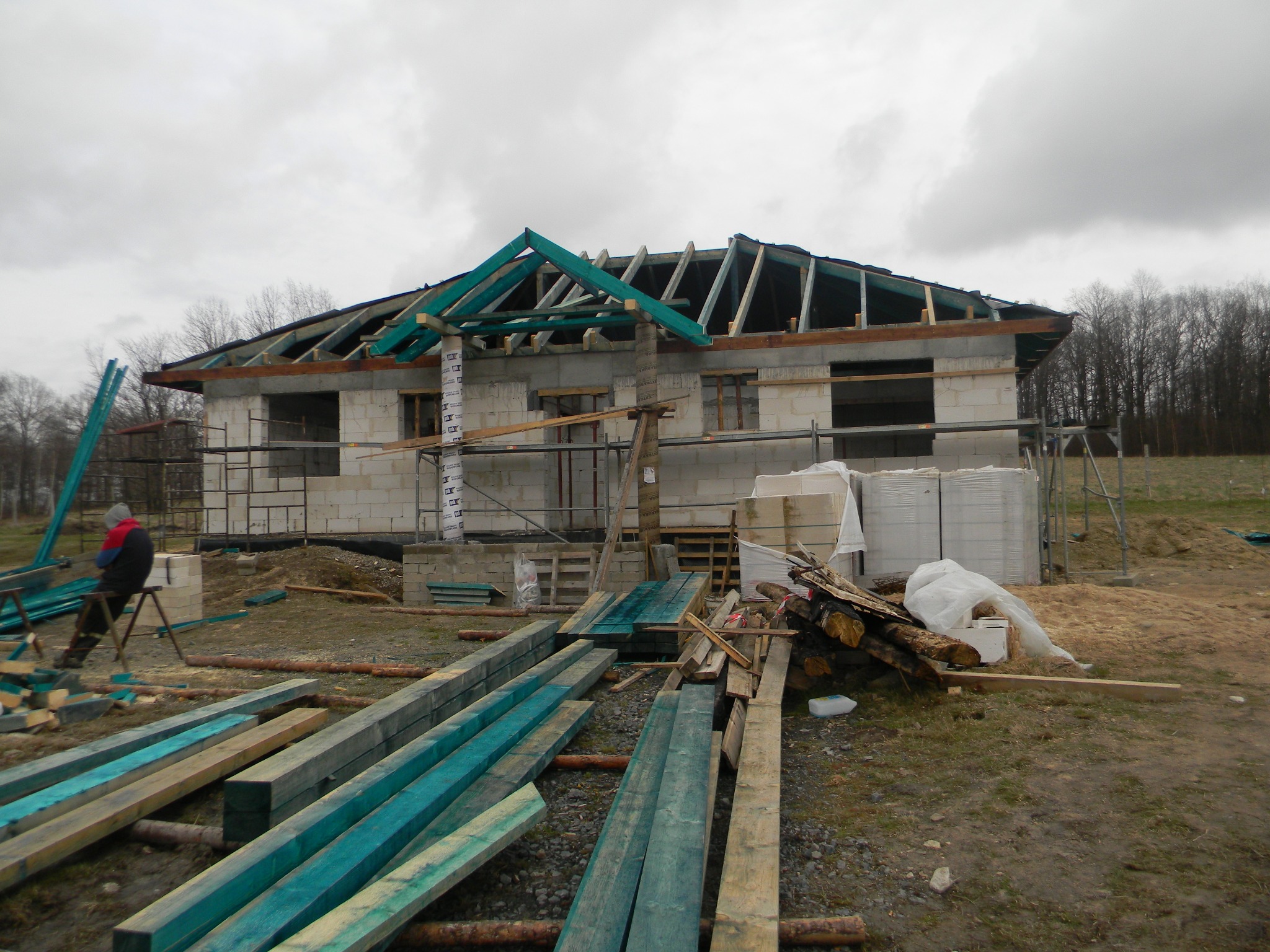 Budowa domu w trakcie realizacji, widoczne ściany z bloczków, drewniana konstrukcja dachu pomalowana na zielono, rusztowania i materiały budowlane na placu budowy.