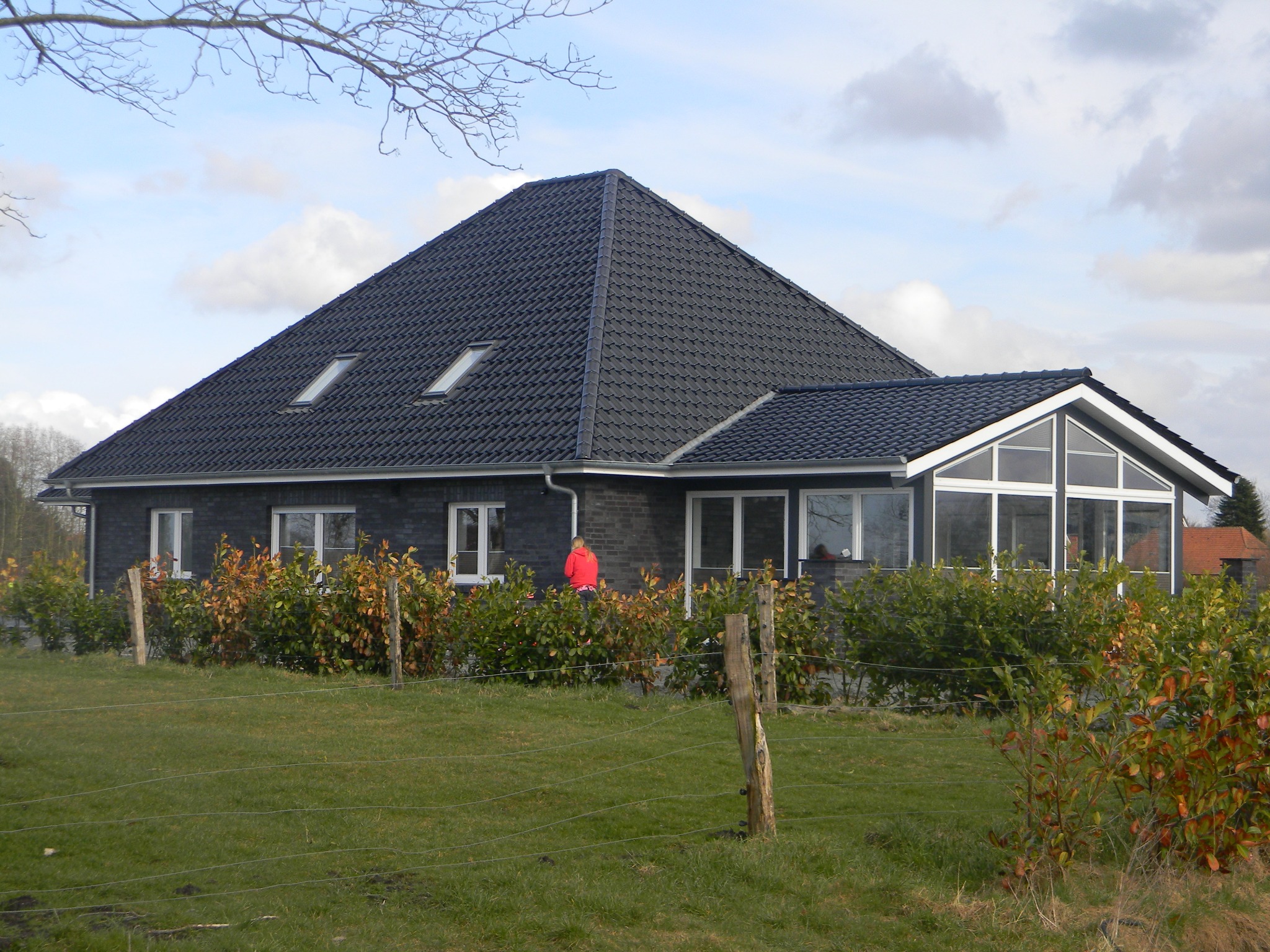 Nowoczesny dom jednorodzinny z ciemną dachówką, lukarnami i przeszkloną werandą, otoczony zielenią i niskim ogrodzeniem.