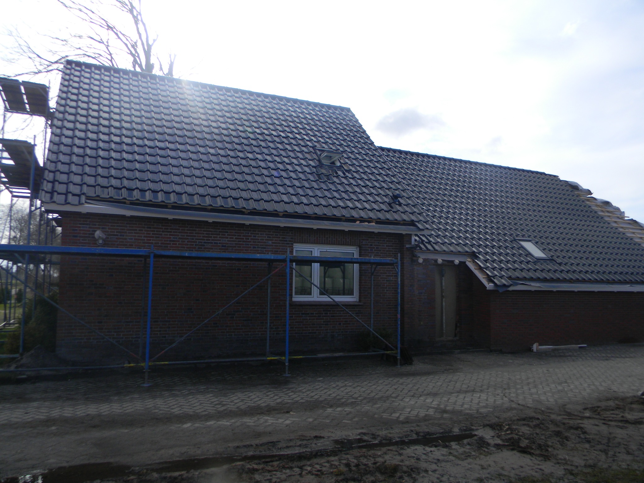 Częściowo wymienione, ciemne dachówki na dachu domu z czerwonej cegły, widoczne rusztowanie i fragment niezakończonej połaci dachu z lukarną.