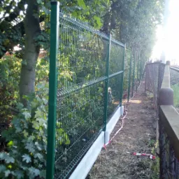 Długi, zielony panelowy system ogrodzeniowy z betonową podmurówką, biegnący wzdłuż torów kolejowych, otoczony zielenią.