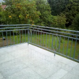 Szara, metalowa balustrada balkonowa z dekoracyjnymi owalami na tle zieleni, widoczne krople deszczu na metalu i płytkach.