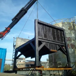 Ciężki dźwig podnosi stalową konstrukcję hali na tle zbiornika przemysłowego i błękitnego nieba.