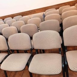 Szałek Krzysztof Szałkowski - Rząd krzeseł z beżową tapicerką i czarnymi metalowymi ramami, ustawionych rzędami na brązowej podłodze, widok z boku pod kątem.