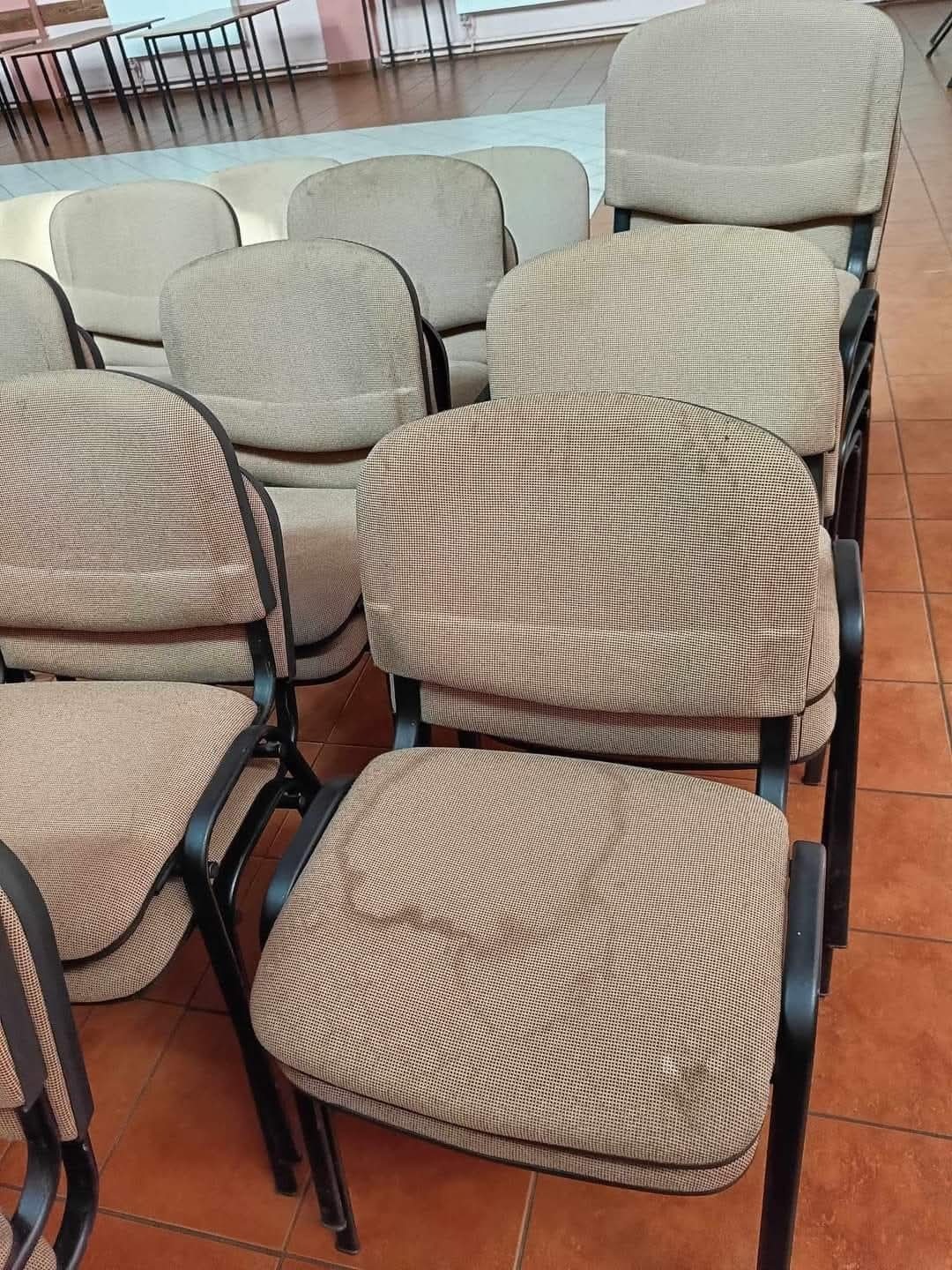 Stos krzeseł biurowych z brązową tapicerką, widoczne plamy na jednym siedzisku, ustawione na pomarańczowej podłodze z płytek.