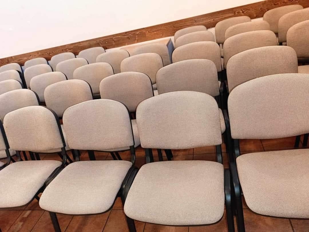 Rząd krzeseł z beżową tapicerką i czarnymi metalowymi ramami, ustawionych rzędami na brązowej podłodze, widok z boku pod kątem.