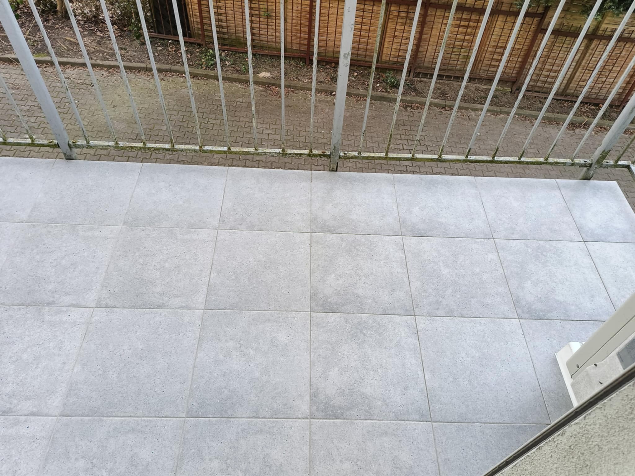 Szare płytki na balkonie w Wolinie, widok z góry, metalowa balustrada z delikatną zielenią u podstawy, w tle brązowe ogrodzenie.