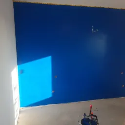 Świeżo pomalowana na niebiesko ściana z widocznym odbiciem światła, zabezpieczona taśmą malarską przy suficie, z wiadrem farby i wałkiem malarskim na podłodze.