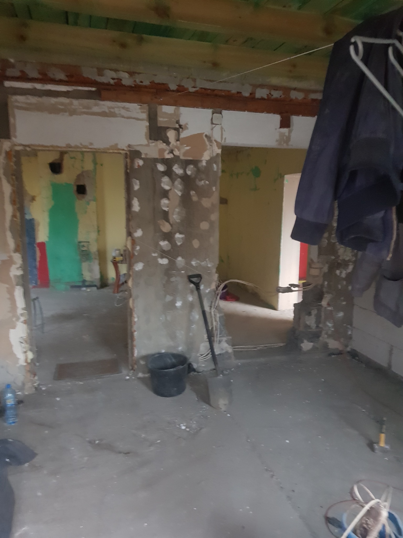 Wnętrze w trakcie demontażu z odsłoniętymi ścianami, widoczne ślady po instalacjach, wiadro, łopata i ubranie robocze wiszące na ścianie.
