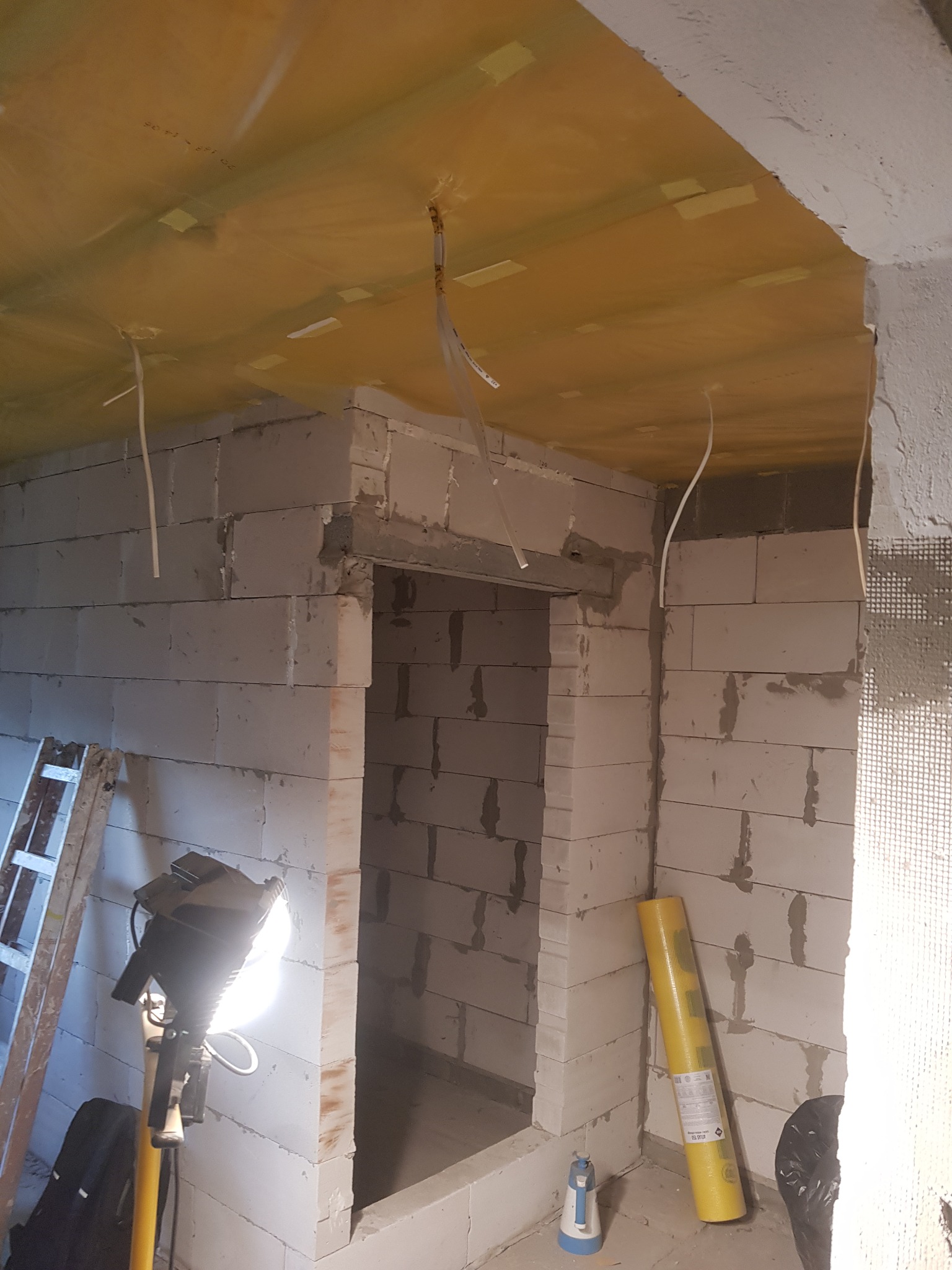 Wnętrze w trakcie budowy: surowy mur z bloczków, żółta folia na suficie przyklejona taśmą, wiszące kable, drabina i lampa budowlana oświetlająca przestrzeń.