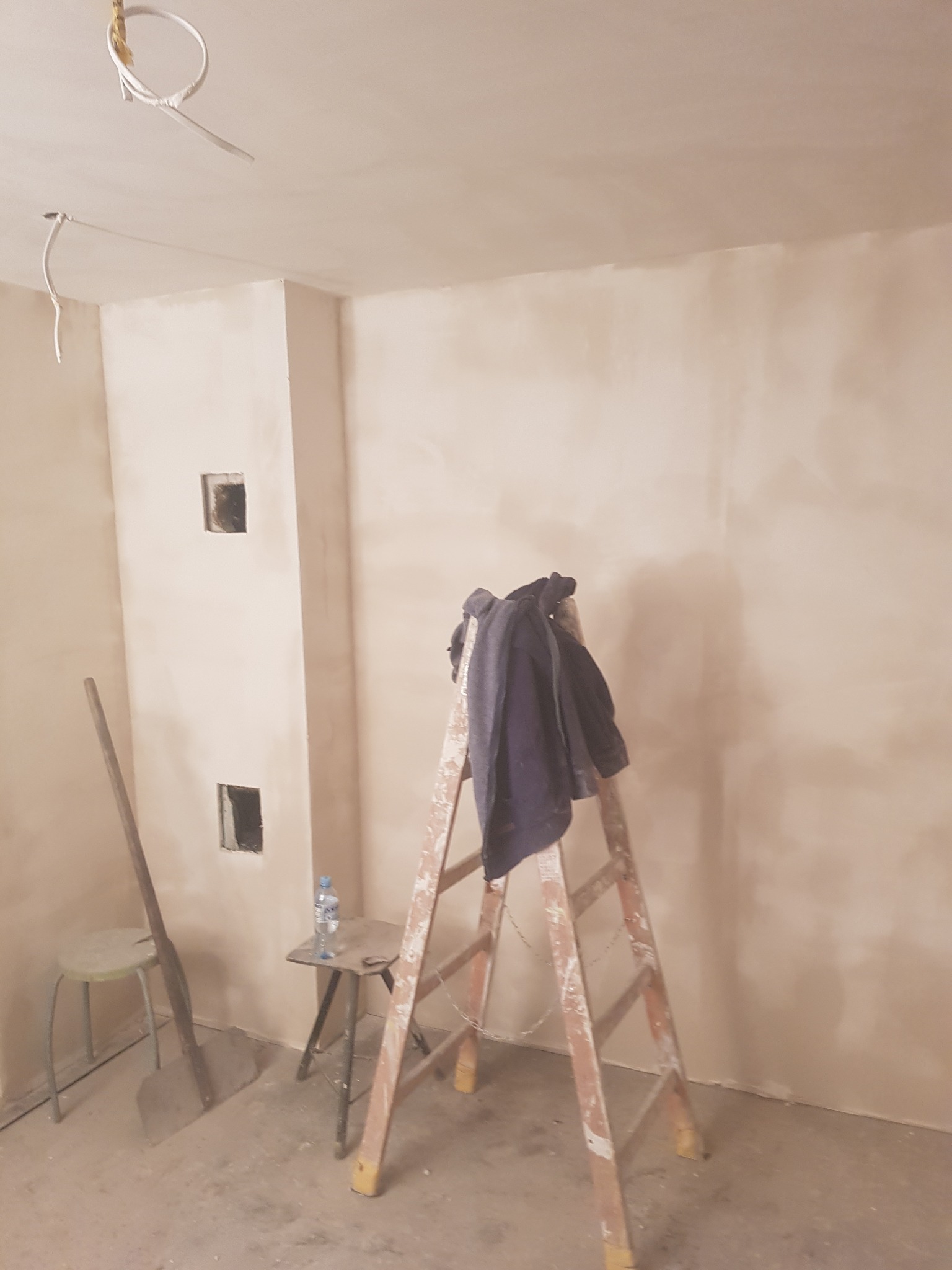 Pokój w trakcie prac wykończeniowych: świeżo położona gładź szpachlowa na ścianach, widoczne otwory na instalacje, drabina z roboczą bluzą, szpachelka i stołek.