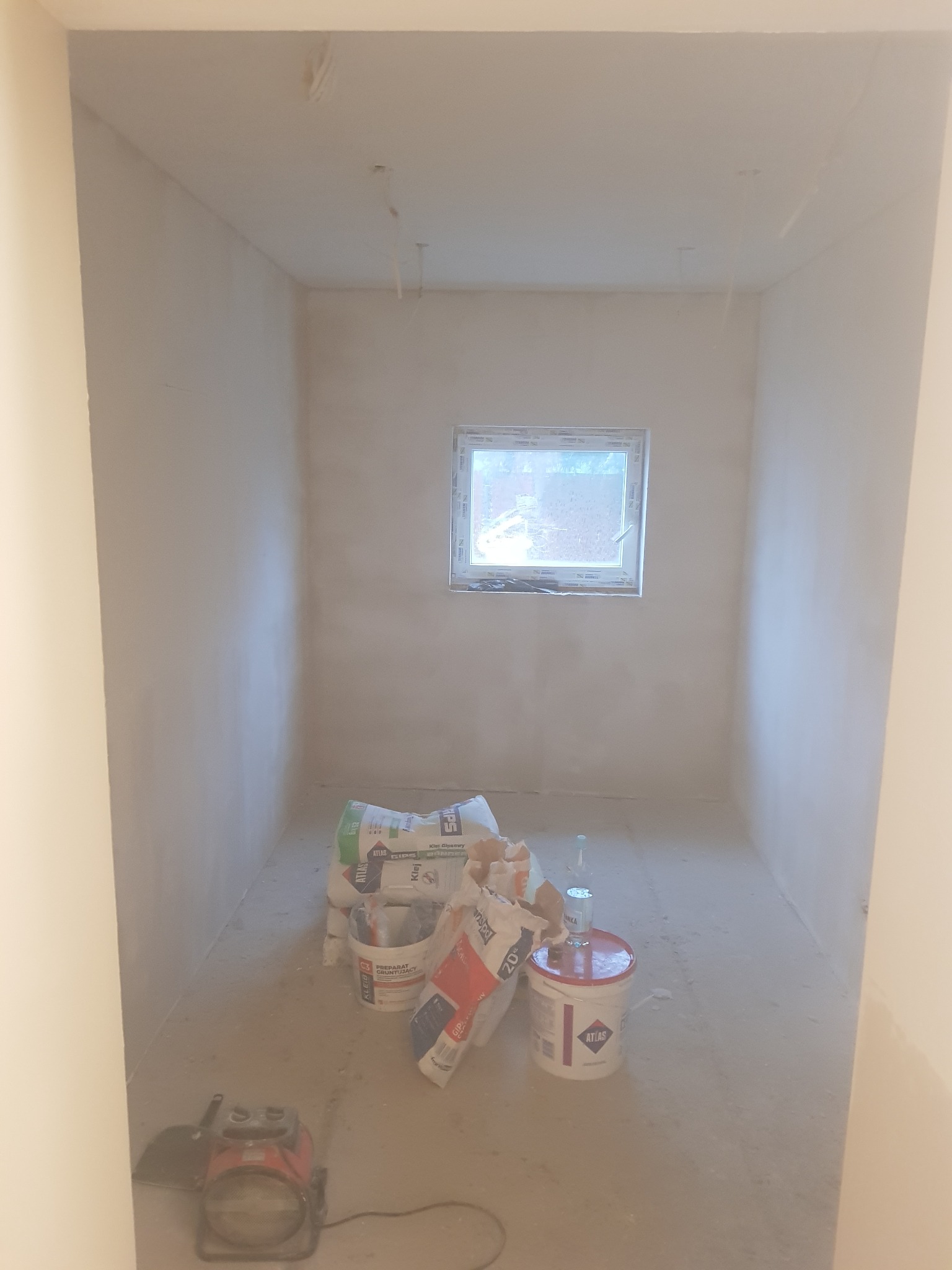 Przygotowane do malowania pomieszczenie z workami gipsu, wiadrami z farbą i grzejnikiem elektrycznym na podłodze.