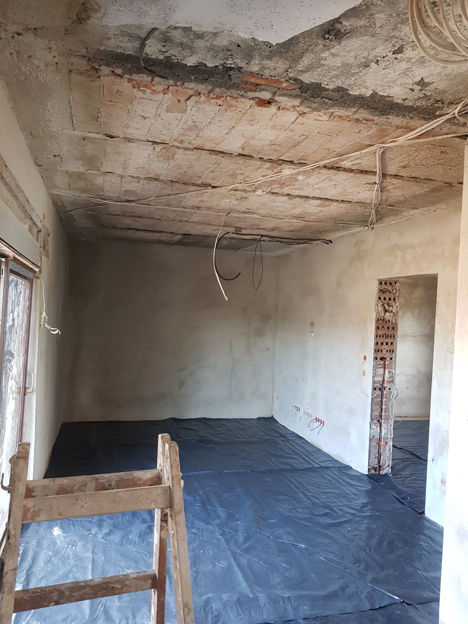 Wnętrze pokoju w trakcie remontu: surowy ceglany strop, otynkowane ściany z widoczną instalacją elektryczną, podłoga zabezpieczona czarną folią, drewniana drabina w rogu pomieszczenia.
