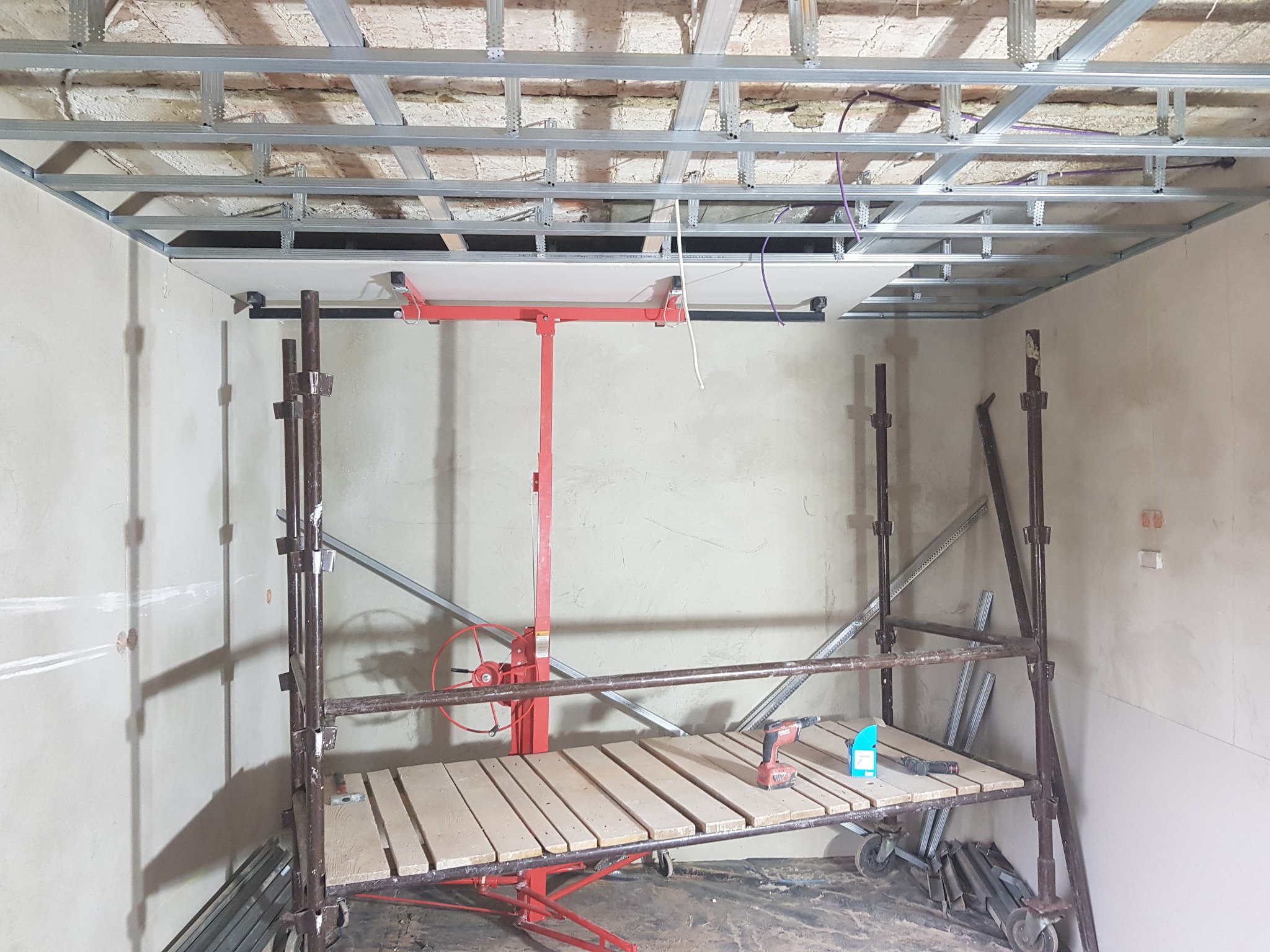 Montaż płyty gipsowo-kartonowej na metalowym stelażu sufitowym, widoczny podnośnik do płyt oraz rusztowanie z wiertarko-wkrętarką na podeście.
