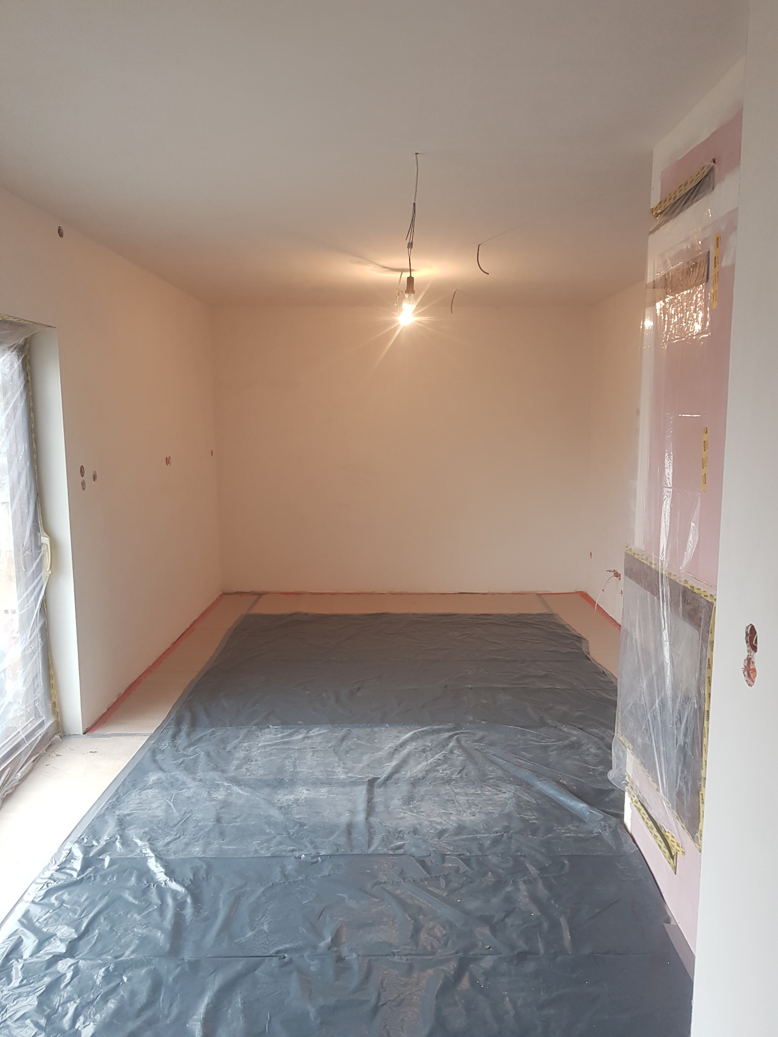 Pokój przygotowany do malowania: podłoga zabezpieczona folią, ściany zagruntowane, widoczne gniazdka elektryczne, prowizoryczne oświetlenie żarówką.