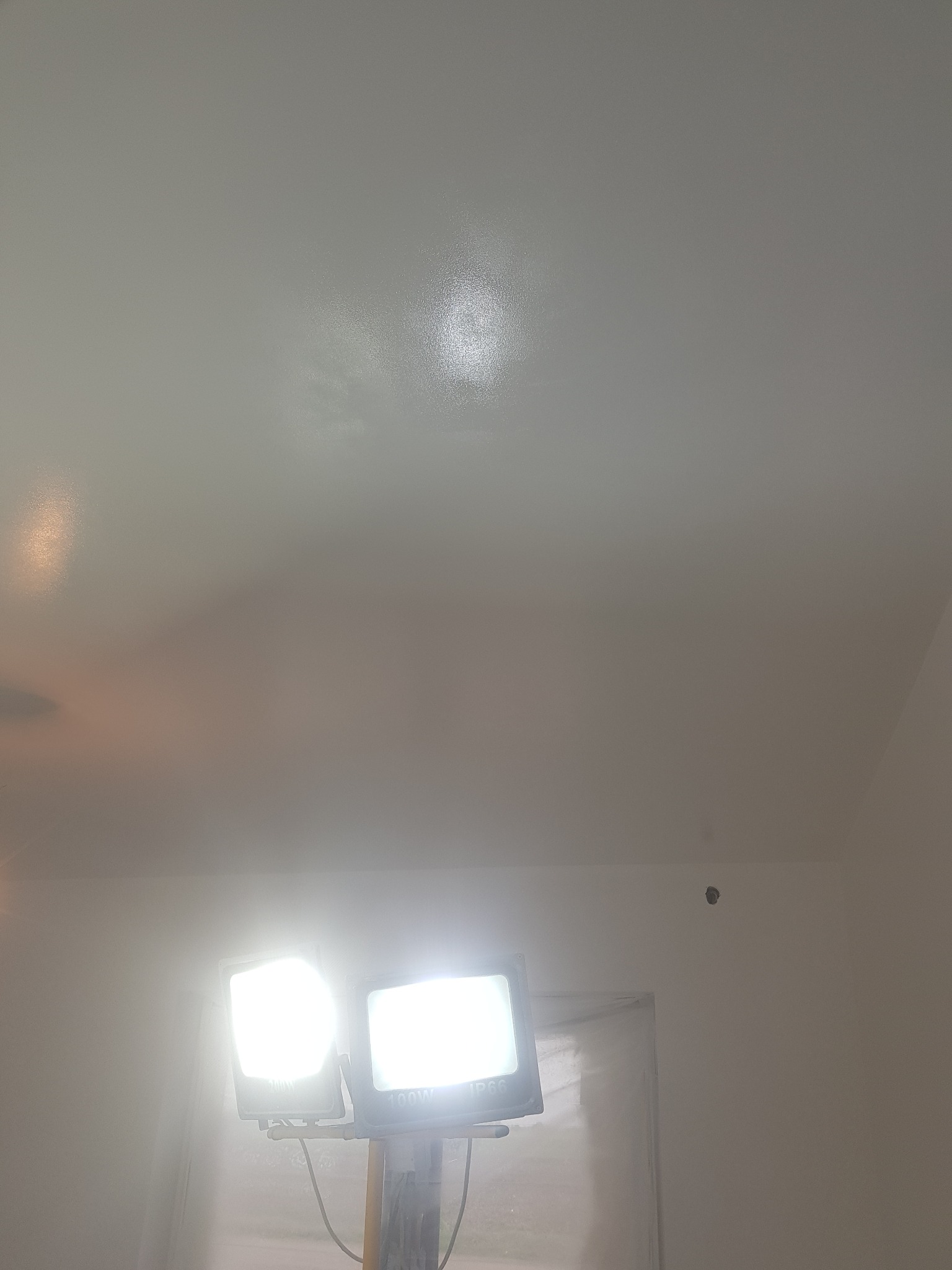 Świeżo pomalowany biały sufit oświetlony dwoma lampami halogenowymi na statywie, widoczne niedoskonałości w fakturze farby.