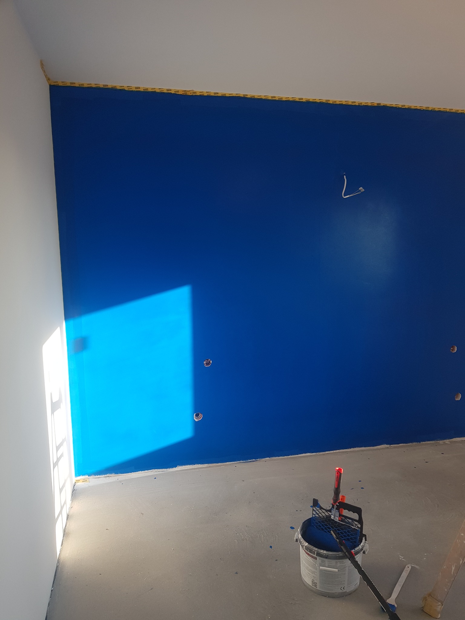 Świeżo pomalowana na niebiesko ściana z widocznym odbiciem światła, zabezpieczona taśmą malarską przy suficie, z wiadrem farby i wałkiem malarskim na podłodze.
