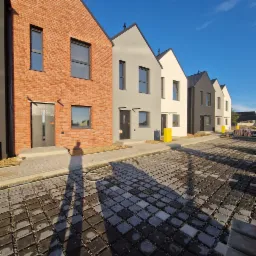 Nowe domy w zabudowie szeregowej o różnej kolorystyce elewacji, z kostką brukową na podjeździe, w słoneczny dzień. Widoczny cień osoby.