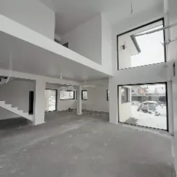 Wnętrze domu w trakcie wykańczania: świeżo położone gładzie na ścianach i suficie, surowa posadzka, duże okna z czarnymi ramami, widoczne instalacje elektryczne przed finalnym wykończeniem.