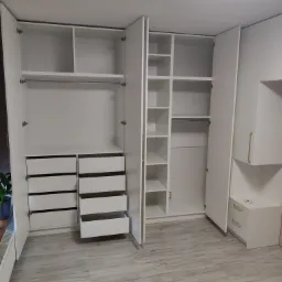 Wnętrze garderoby z białymi meblami na wymiar, widoczne otwarte drzwi szaf, półki i szuflady, jasna podłoga imitująca drewno.