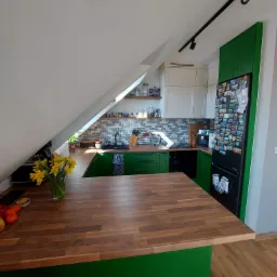 Kuchnia w mieszkaniu na poddaszu z zielonymi szafkami, drewnianym blatem i czarną lodówką obklejoną magnesami, oświetlona światłem dziennym wpadającym przez okno dachowe.