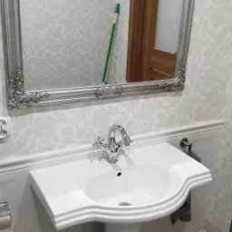 Umywalka na postumencie z chromowaną baterią i ozdobnym lustrem w srebrnej ramie w łazience z tapetą w kwiaty; w lustrze odbija się zielony mop.