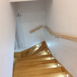 Drewniane schody z podświetleniem LED wzdłuż krawędzi stopni i drewnianą balustradą na białej ścianie, widok z góry.