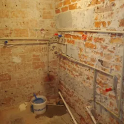 Surowe wnętrze z odsłoniętą cegłą, prezentujące instalację wodną z rurami PCV, zaworami z czerwonymi i niebieskimi pokrętłami, oraz wiadro z niebieską szufelką w rogu.