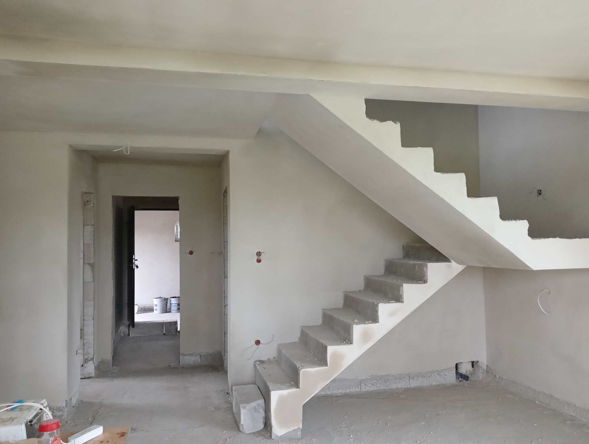 Wnętrze w trakcie prac wykończeniowych: surowe schody betonowe, świeżo otynkowane ściany z widocznymi puszkami elektrycznymi i otworem drzwiowym prowadzącym do kolejnego pomieszczenia z wiadrami farb.