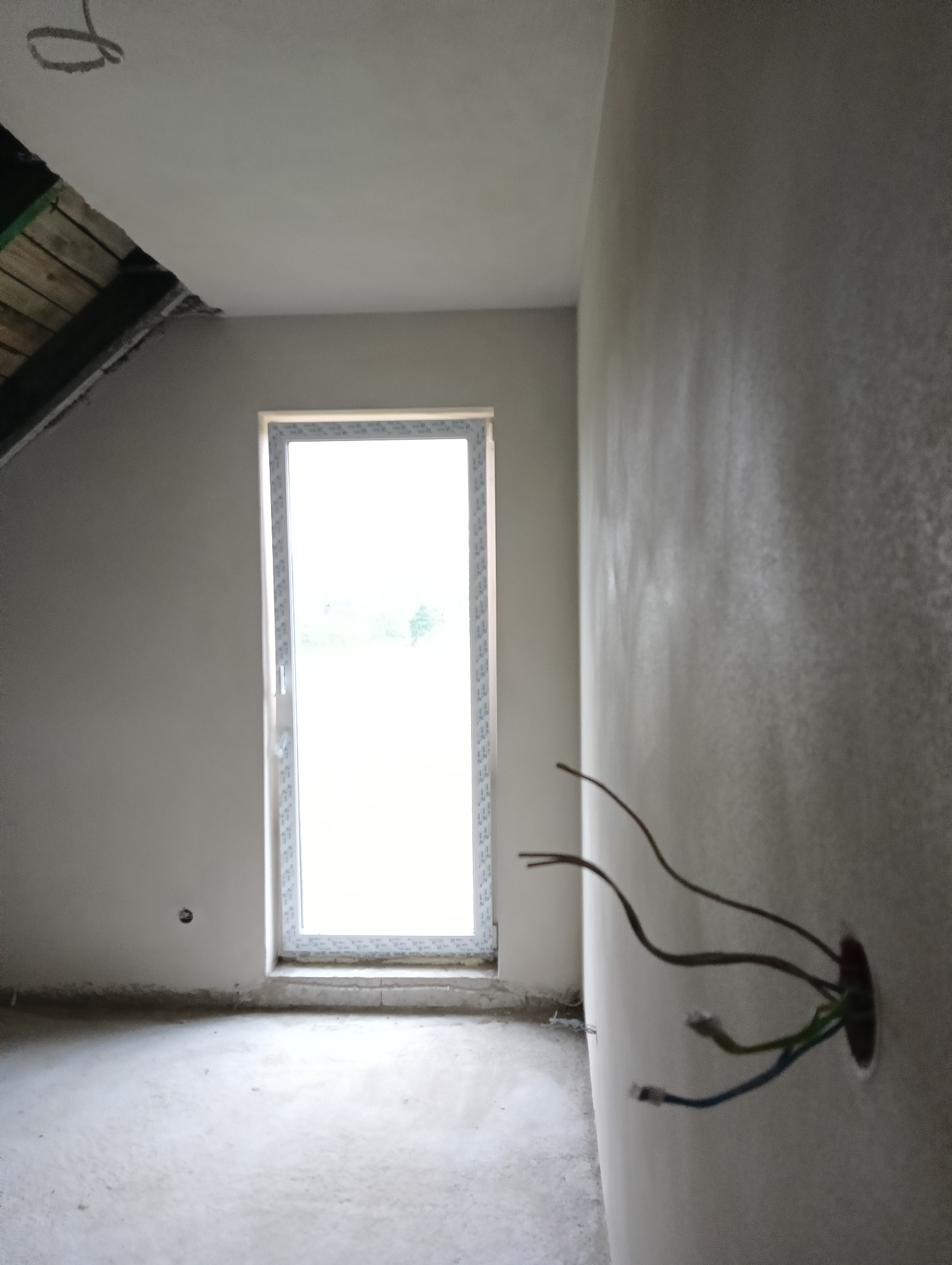 Pokój na poddaszu w trakcie prac wykończeniowych, ściany pokryte świeżym tynkiem, widoczne wystające przewody elektryczne, okno w białej ramie.
