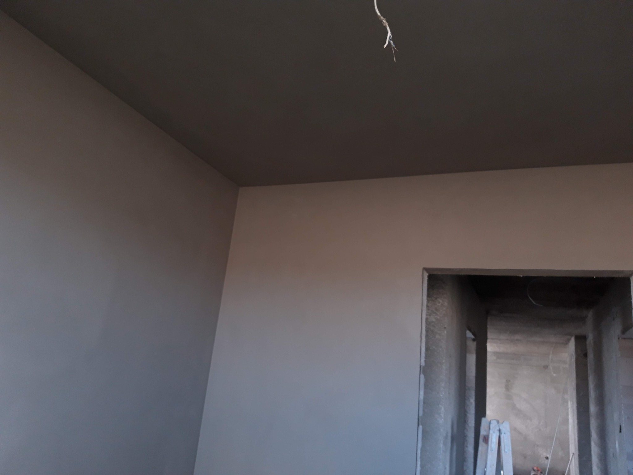 Surowe wnętrze w trakcie prac wykończeniowych: gładkie, jasne ściany i ciemniejszy sufit, widoczne kable zwisające z sufitu, w tle otwór drzwiowy z surowym betonem i fragment drabiny.