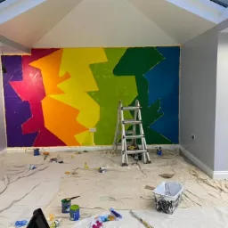 Wnętrze pokoju z tęczową, geometryczną malowanką na ścianie, drabina oparta o ścianę, podłoga zabezpieczona folią malarską, widoczne pędzle i pojemniki z farbami.