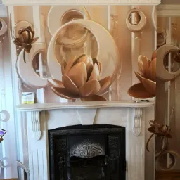 Ściana z wzorzystą tapetą w odcieniach beżu i złota, przedstawiająca motywy kwiatowe i geometryczne, nad marmurowym kominkiem z ozdobnym metalowym wkładem.