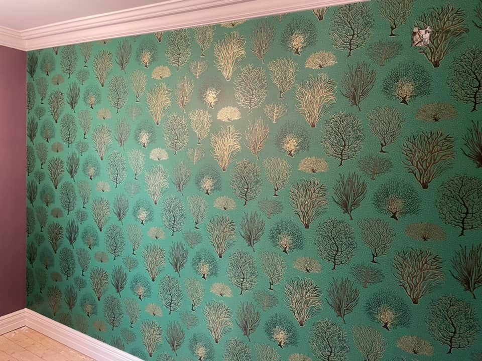 Ściana pokryta zieloną tapetą w motywy drzew, widoczne łączenia tapety przy suficie i w rogu pokoju.