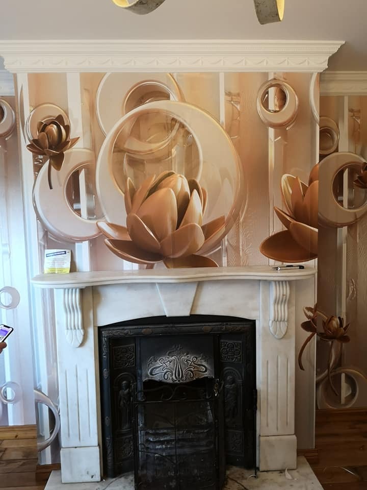 Ściana z wzorzystą tapetą w odcieniach beżu i złota, przedstawiająca motywy kwiatowe i geometryczne, nad marmurowym kominkiem z ozdobnym metalowym wkładem.