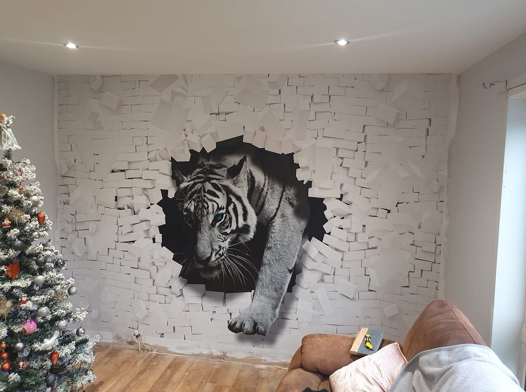 Fototapeta 3D z motywem tygrysa wychodzącego ze ściany z białej cegły, po lewej choinka, w tle widoczne fragmenty sofy.