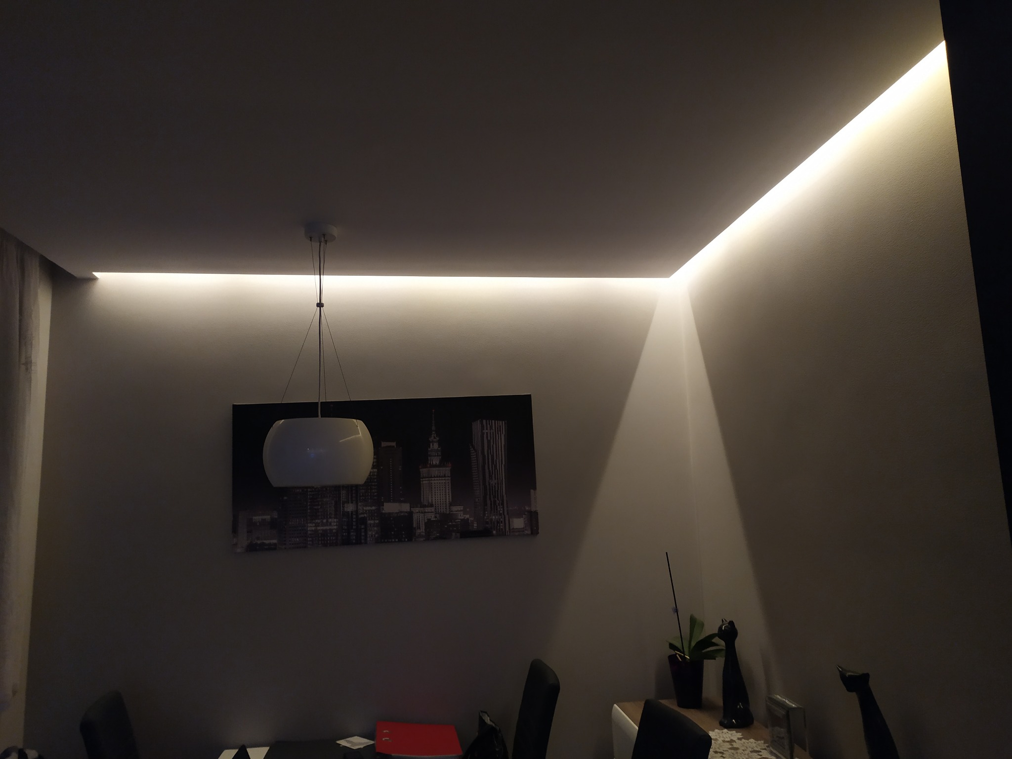 Nowoczesne oświetlenie sufitowe LED w salonie z obrazem panoramy miasta na ścianie, widoczne lampy wiszące i dekoracje.