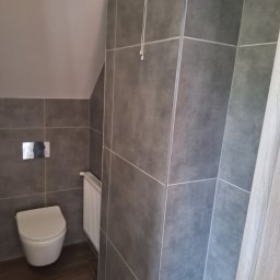 usługi remontowo budowlane - Nowoczesna łazienka z szarymi płytkami, podwieszaną toaletą i grzejnikiem drabinkowym na tle skośnego sufitu.