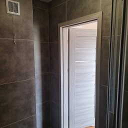 usługi remontowo budowlane - Nowoczesna łazienka z szarymi płytkami, drewnopodobną podłogą i jasnoszarymi drzwiami z poziomymi frezami, widoczny fragment kabiny prysznicowej i kratka wentylacyjna.