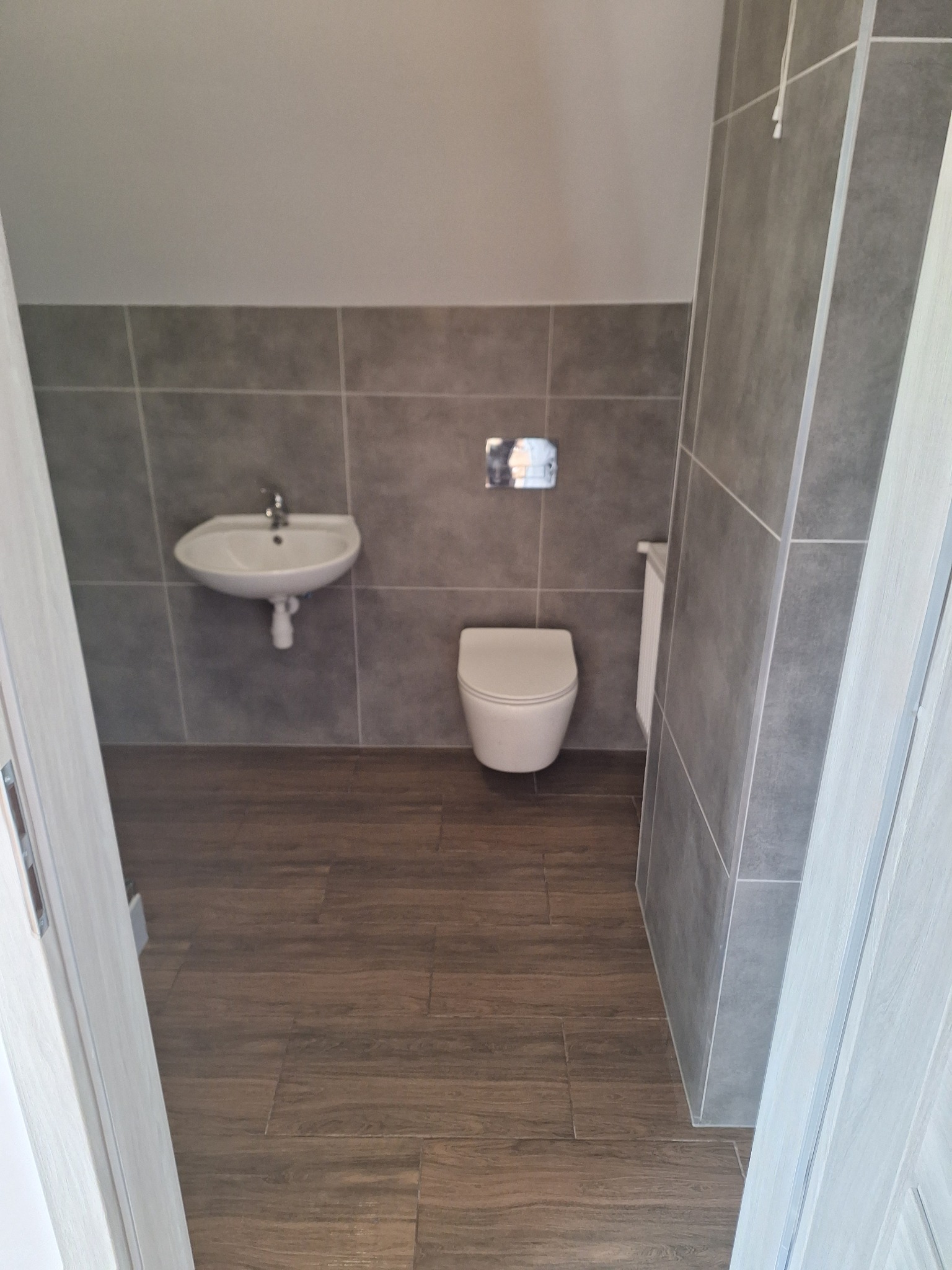 Nowoczesna łazienka z szarymi płytkami na ścianach, brązowymi płytkami na podłodze, umywalką i podwieszaną toaletą, widok zza uchylonych drzwi.