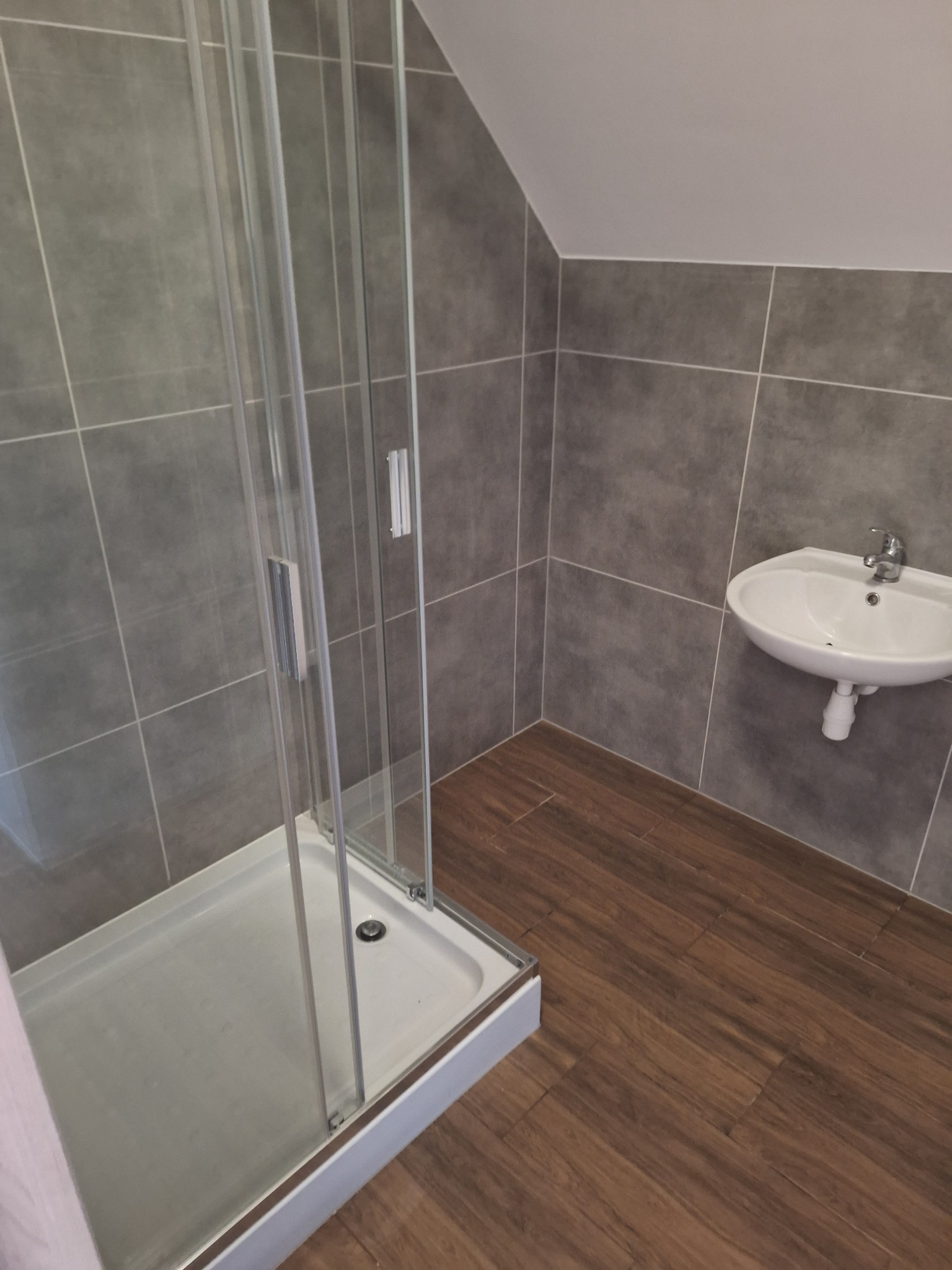 Nowoczesna łazienka z szarą glazurą, kabiną prysznicową z przezroczystego szkła i drewnopodobną podłogą, uzupełniona białą umywalką.